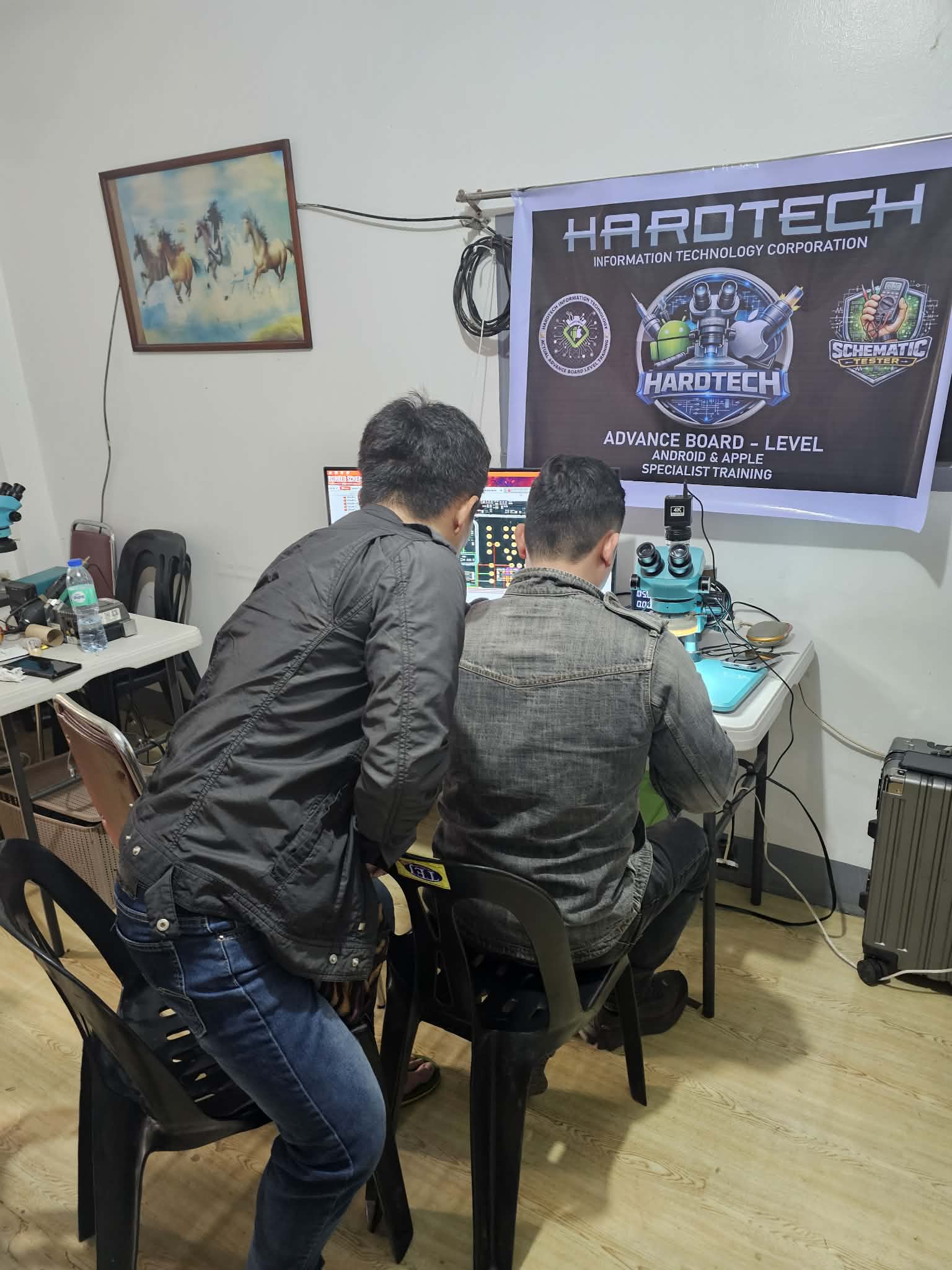 Two trainees observing a board-repair workstation beneath a HardTech banner.
